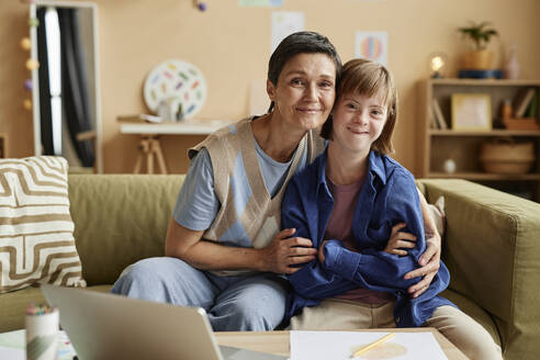 Lächelnde Mutter und ihre Tochter mit Down-Syndrom sitzen auf dem Sofa - KPEF00412