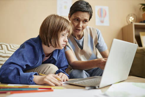 Mother explaining lesson on laptop to daughter with Down syndrome at home - KPEF00414