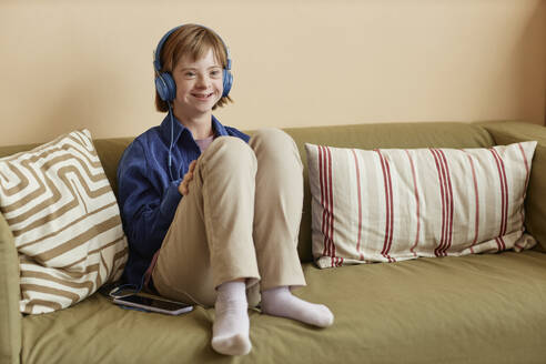 Lächelndes Mädchen mit Down-Syndrom, das Kopfhörer trägt und zu Hause auf dem Sofa sitzt - KPEF00434