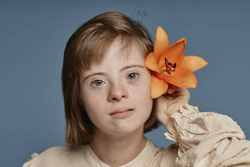 Girl with down syndrome holding orange orchid behind ear against blue background - KPEF00461