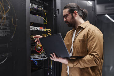 Ingenieur hält Laptop und prüft Kabel im Serverraum - KPEF00472