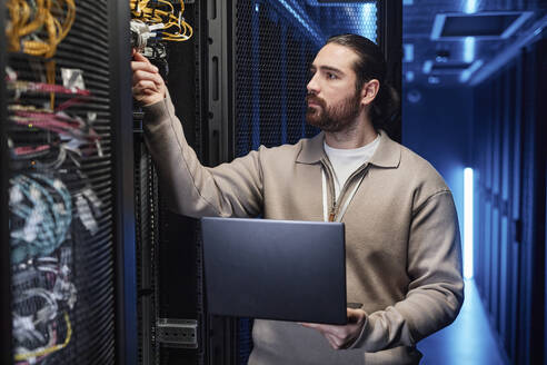 IT-Fachmann hält Laptop und prüft Kabel im Serverraum - KPEF00500