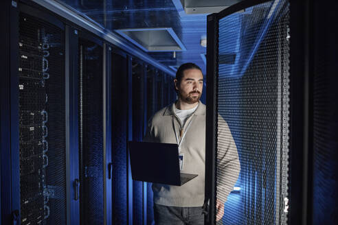 Engineer holding laptop and examining supercomputer in server room - KPEF00510