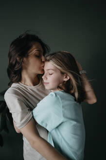 Loving mother hugging and kissing forehead of daughter at home - ANAF02523