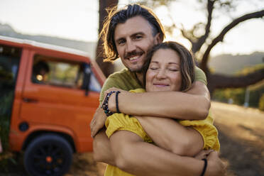 Smiling man embracing female friend standing by caravan - ANNF00672