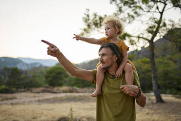 Mann zeigt auf seine Tochter und trägt sie auf den Schultern vor einem klaren Himmel - ANNF00722