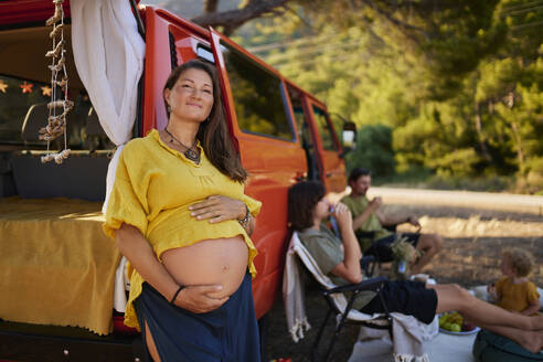 Lächelnde schwangere Frau, die sich an den Wohnwagen einer im Park sitzenden Familie lehnt - ANNF00769