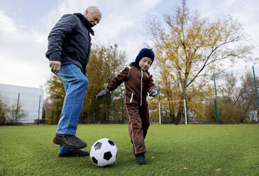 Älterer Mann und Enkel spielen mit Ball auf einem Fußballplatz - MBLF00168