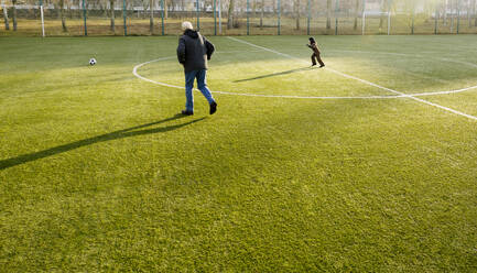 Großvater und Enkel laufen auf dem Fußballplatz dem Ball hinterher - MBLF00175