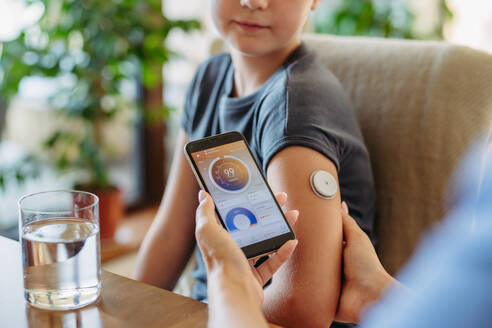 Mädchen mit Diabetes, das zu Hause mit einem kontinuierlichen Glukosemessgerät den Blutzuckerspiegel kontrolliert. Die Mutter des Mädchens verbindet das CGM mit einem Smartphone, um den Blutzuckerspiegel in Echtzeit zu überwachen. - HPIF31353