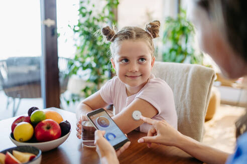 Mädchen mit Diabetes, das zu Hause mit einem kontinuierlichen Glukosemessgerät den Blutzuckerspiegel kontrolliert. Die Mutter des Mädchens verbindet das CGM mit einem Smartphone, um den Blutzuckerspiegel in Echtzeit zu überwachen. - HPIF31356
