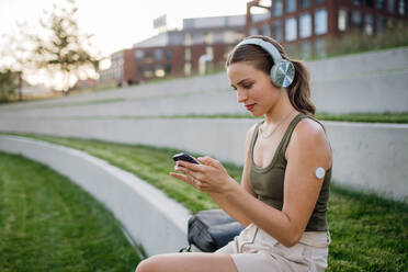 Diabetikerin mit kontinuierlichem Blutzuckermessgerät, die im Park sitzt und soziale Medien auf ihrem Smartphone anschaut: Das CGM-Gerät erleichtert das Leben der Schülerin. - HPIF31389