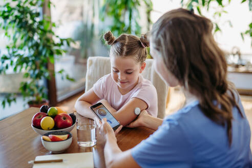 Mädchen mit Diabetes, das zu Hause mit einem kontinuierlichen Glukosemessgerät den Blutzuckerspiegel kontrolliert. Die Mutter des Mädchens verbindet das CGM mit einem Smartphone, um den Blutzuckerspiegel in Echtzeit zu überwachen. - HPIF31398