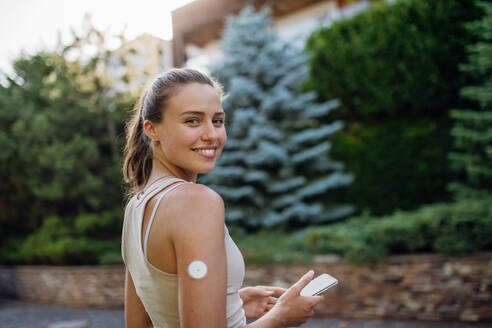 Schöne Diabetikerin, die sich auf ein Outdoor-Training in der Stadt vorbereitet. Junge Frau mit CGM, die ihren Blutzuckerspiegel vor dem Training überprüft. Konzept von Bewegung und Diabetes. - HPIF31414