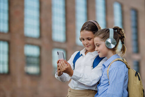 Zwei Schülerinnen checken vor der Schule soziale Medien auf ihrem Smartphone, während Freunde mit Kopfhörern ein Video ansehen. - HPIF31433