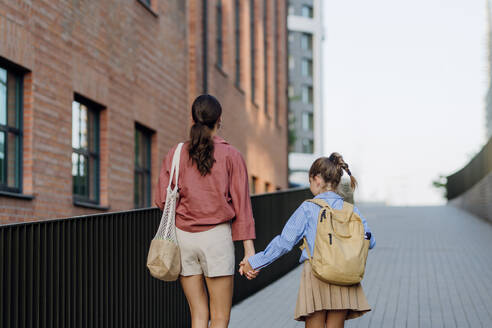 Rückansicht einer Mutter und eines kleinen Mädchens, die sich an den Händen halten und zum ersten Mal zur Schule gehen. Das kleine Schulmädchen geht in die erste Klasse. Konzept der Einschulung. - HPIF31434