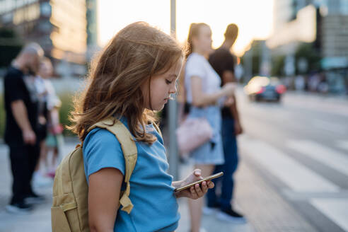 Seitenansicht eines Mädchens, das auf dem Fußgängerüberweg wartet und auf dem Weg zur Schule auf ihrem Smartphone scrollt. Konzept des Schulanfangs. - HPIF31446