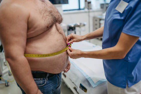 Ärztin misst die Taille eines übergewichtigen Patienten mit einem Maßband. Übergewicht beeinträchtigt die Gesundheit von Männern mittleren Alters. Konzept der Gesundheitsrisiken von Übergewicht und Adipositas. - HPIF31448