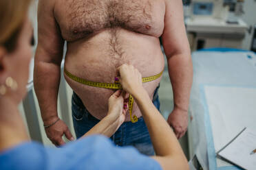 Ärztin misst die Taille eines übergewichtigen Patienten mit einem Maßband. Übergewicht beeinträchtigt die Gesundheit von Männern mittleren Alters. Konzept der Gesundheitsrisiken von Übergewicht und Adipositas. - HPIF31449
