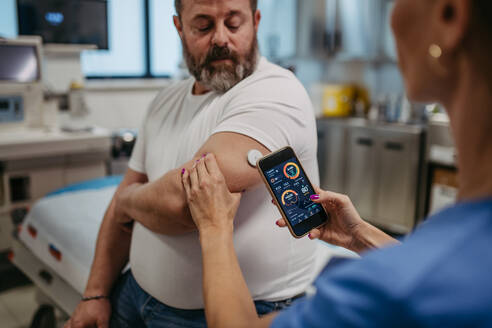 Doctor connecting patient's continuous glucose monitor with smartphone, to check his blood sugar level in real time. Obese, overweight man is at risk of developing type 2 diabetes. Concept of health risks of overwight and obesity. - HPIF31477