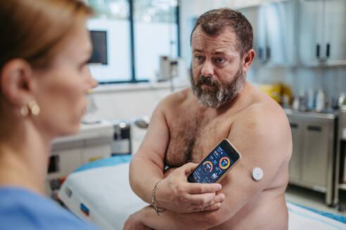 Der Arzt verbindet das kontinuierliche Glukosemessgerät des Patienten mit dem Smartphone, um den Blutzuckerspiegel in Echtzeit zu überprüfen. Ein übergewichtiger Mann ist gefährdet, an Typ-2-Diabetes zu erkranken. Konzept der Gesundheitsrisiken von Übergewicht und Adipositas. - HPIF31480