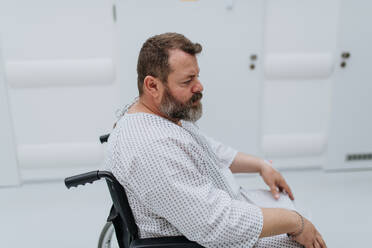 Seitenansicht des Mannes im Rollstuhl. Übergewichtiger Patient im Krankenhauskittel, der auf eine medizinische Untersuchung im Krankenhaus wartet und sich ängstlich fühlt. - HPIF31497