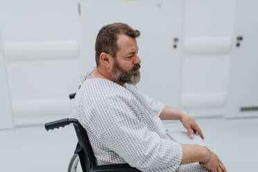 Side view of the man in wheelchair. Overweight patient in hospital gown waiting for medical examination in hospital, feeling anxious. - HPIF31497