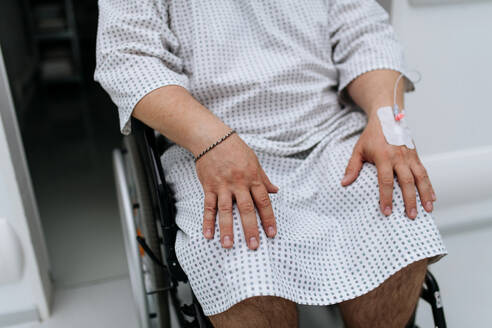 Nahaufnahme eines Mannes im Rollstuhl mit einer Infusionskanüle in der Hand. Übergewichtiger Patient im Krankenhauskittel wartet auf eine medizinische Untersuchung im Krankenhaus. - HPIF31498