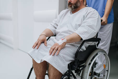 Krankenpfleger, der einen Patienten im Rollstuhl über einen Krankenhausflur schiebt, der auf eine medizinische Untersuchung wartet. Der übergewichtige Patient fühlt sich ängstlich und hat gesundheitliche Bedenken. - HPIF31499