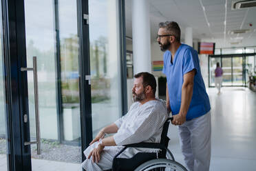 Krankenpfleger, der einen Patienten im Rollstuhl über einen Krankenhausflur schiebt. Der übergewichtige Patient fühlt sich unruhig und hat gesundheitliche Bedenken. - HPIF31504