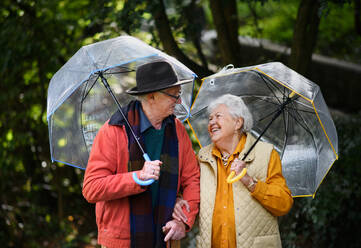 Glückliches älteres Paar, das gemeinsam mit Regenschirmen in einem Park spazieren geht. - HPIF31622