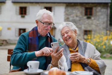 Happy senior couple in park cafe resting after walk, having a coffee and looking at smartphone. - HPIF31643