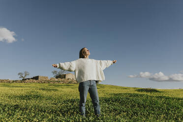 Carefree young woman with arms outstretched standing on grass under sky - DMGF01124