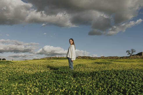 Lächelnde junge Frau unter bewölktem Himmel auf einem Feld stehend - DMGF01127