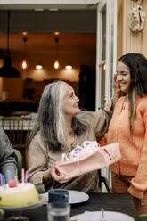 Smiling granddaughter giving birthday present to grandmother at patio - MASF40582