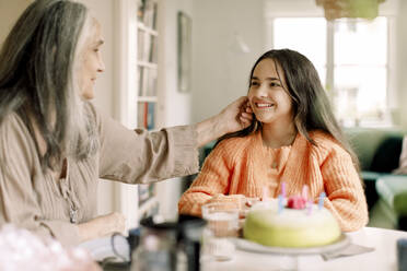 Senior woman touching cheek of granddaughter sitting with birthday cake at dining table - MASF40593