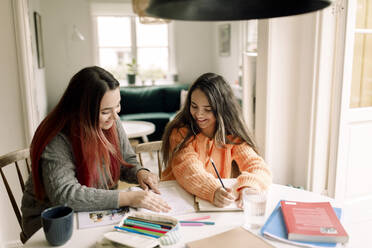 Happy woman assisting daughter in homework while sitting at table - MASF40620