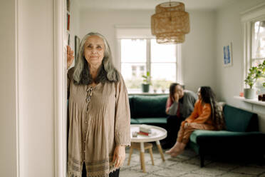 Porträt einer älteren Frau, die sich an die Wand lehnt, während ihre Familie auf dem Sofa im Hintergrund sitzt - MASF40632