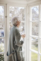 Contemplative senior man holding coffee cup while looking through window at home - MASF40655