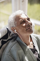 Lächelnder älterer Mann, der zu Hause im Sonnenlicht Musik über In-Ear-Kopfhörer hört - MASF40657