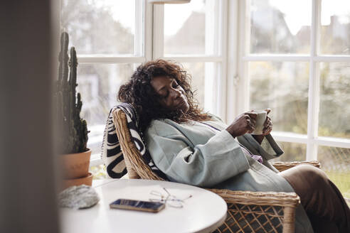 Mature woman holding coffee cup while relaxing on chair at home - MASF40659