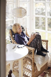 Senior woman with coffee cup looking away while relaxing on chair at home - MASF40668