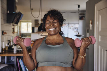 Mature woman clenching teeth while exercising with dumbbells at home - MASF40677