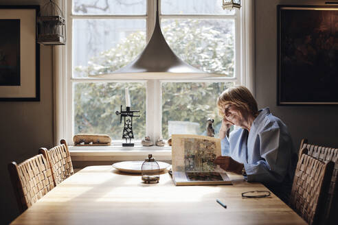 Smiling senior woman looking at picture book on dining table at home - MASF40681
