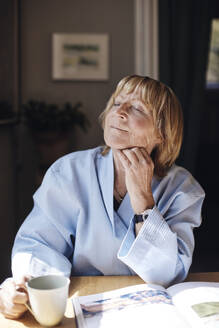 Senior woman with hand on chin holding coffee cup while relaxing at table - MASF40682