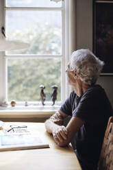 Senior man looking through window while sitting at home - MASF40687