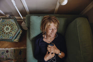 Lächelnde ältere Frau mit Smartphone hört Musik auf dem Sofa zu Hause - MASF40699
