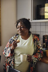 Mature woman with eyes closed having cookie and coffee in kitchen at home - MASF40700