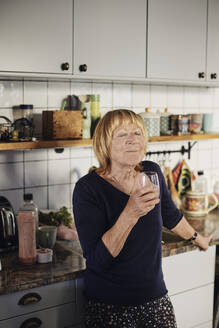 Blond senior woman with eyes closed holding drinking glass in kitchen at home - MASF40703