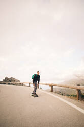 Ein junger Mann gleitet auf seinem Skateboard eine Straße entlang, mit einer hölzernen Leitplanke und einem atemberaubenden Himmel im Hintergrund - MASF40751